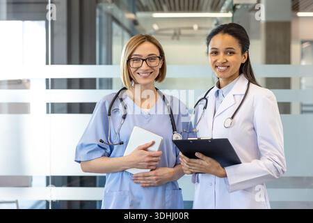 Due diverse donne professionisti medici infermiera in scrub blu con tablet e medico in camice da laboratorio con appunti sorridente insieme, che rappresentano lavoro di squadra, fiducia e cura centrata sul paziente Foto Stock