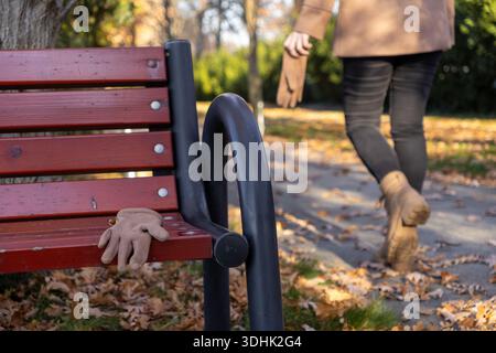 Primo piano di un guanto marrone chiaro su una panchina rossa in legno del parco, con una persona sfocata che si allontana sullo sfondo, incarnando i temi di una passeggiata autunnale Foto Stock