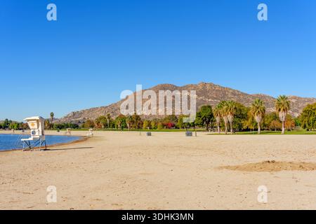 Il bagnino bianco sorge su un'ampia spiaggia sabbiosa che confina con il lago Perris, montagne rocciose e alberi diversi visibili sotto un cielo blu brillante. Foto Stock