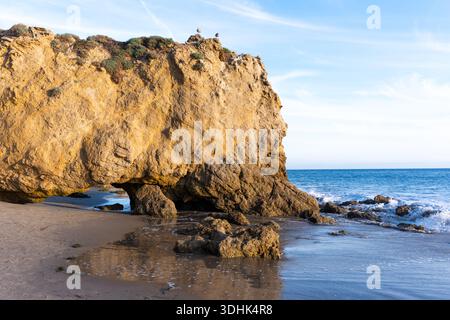 La luce del sole dorata illumina splendidamente un maestoso arco di roccia naturale a El Matador State Beach, con onde spumeggianti che lambiscono dolcemente lo sho sabbioso Foto Stock