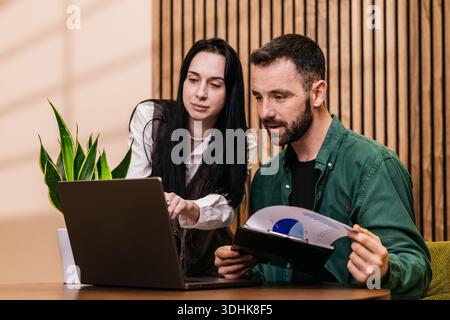 Due professionisti si impegnano in una discussione a un tavolo di legno, con una donna che indica il laptop mentre l'uomo esamina un rapporto, ambiente interno Foto Stock