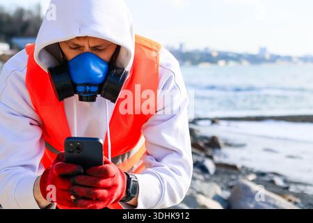 Indossa il respiratore e i guanti utilizzando lo smartphone sulla costa rocciosa con vista sul mare e sugli edifici urbani distanti. Foto Stock