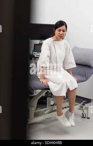 Donna seduta sul tavolo d'esame in clinica che indossa camice ospedaliero e scarpe da ginnastica bianche, monitor nelle vicinanze Foto Stock
