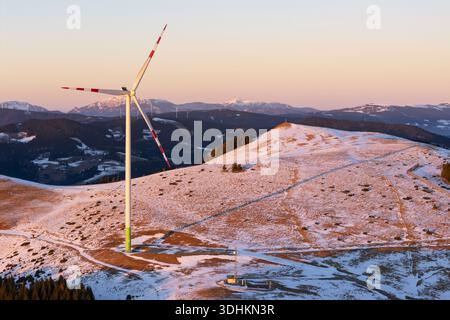 Vista aerea di una sola turbina eolica che si erge su uno sfondo di colline innevate sotto il cielo pastello, Sommeralm, Steiermark, Austria. Foto Stock