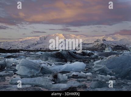 Vista di iceberg luminosi sparsi attraverso la laguna glaciale sotto un cielo di cotone caramellato, con montagne innevate in lontananza, Jökulsárlón, Sveit Foto Stock