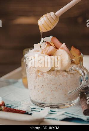 Un vasetto di vetro pieno di cremosa avena notturna condita con banane a fette, fragole, fiocchi di cocco e un filo di miele. Il vaso viene posizionato su un Foto Stock
