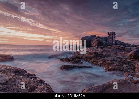 Una tranquilla scena costiera al tramonto, caratterizzata da rocce aspre e calme onde oceaniche. Foto Stock
