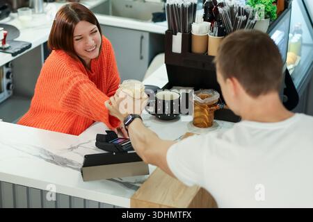 Una donna amichevole con maglione arancione estende la bevanda frizzante al giovane uomo che spillano la carta al bancone del caffè contemporaneo Foto Stock