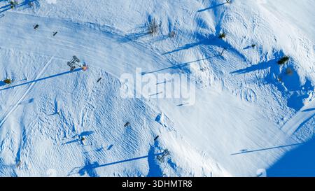 Vista aerea dall'alto verso il basso delle piste da sci alpine con sciatori, lunghe ombre e superficie nevosa strutturata, che illustrano le attività sportive invernali e l'infrazione del resort Foto Stock
