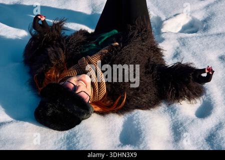 Una donna felice dai capelli rossi con un cappotto di pelliccia e un cappello giace sulla schiena nella neve. Sorride al sole, indossa gli occhiali e si gode l'inverno Foto Stock