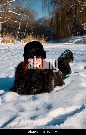 Questa immagine ritrae un'allegra donna dai capelli rossi vestita con un lussuoso cappotto di pelliccia e un cappello coordinato, comodamente sdraiata nella neve incontaminata. La sua gioia Foto Stock