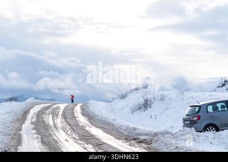 Un veicolo è parcheggiato a lato di una strada innevata e tortuosa. Il paesaggio presenta montagne e un cielo nuvoloso. La neve copre il terreno e le parti della r Foto Stock