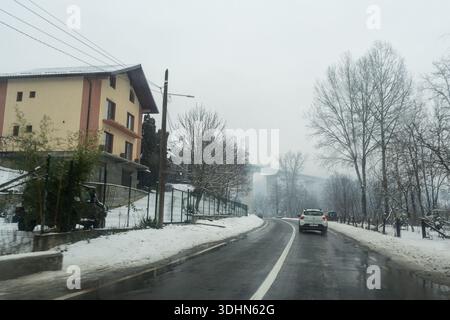 La neve copre il terreno e gli alberi lungo una strada tortuosa. Un'auto bianca passa davanti a diverse case. Un ponte è visibile in lontananza, circondato dalla nebbia Foto Stock