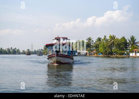Alappuzha, India - 6 marzo 2023: Si vede un traghetto passeggeri che viaggia sulle acque secondarie con palme, case e altre barche visibili lungo la costa sotto un cielo parzialmente nuvoloso. Foto Stock