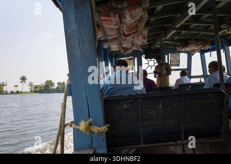 Alappuzha, India - 6 marzo 2023: Diverse persone sono sedute all'interno di un traghetto locale che viaggia nelle acque secondarie, con un membro dell'equipaggio in piedi e attrezzature di sicurezza visibili sopra la testa. Foto Stock