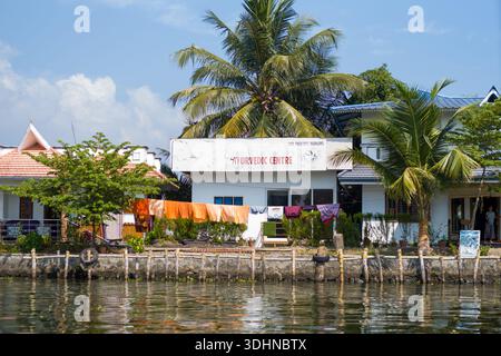 Alappuzha, India - 6 marzo 2023: Un edificio con un cartello con la scritta "Centro Ayurvedico" è visibile sul lungomare, con lavanderia appesa all'esterno, palme e persone visibili vicino all'ingresso sotto un cielo limpido. Foto Stock