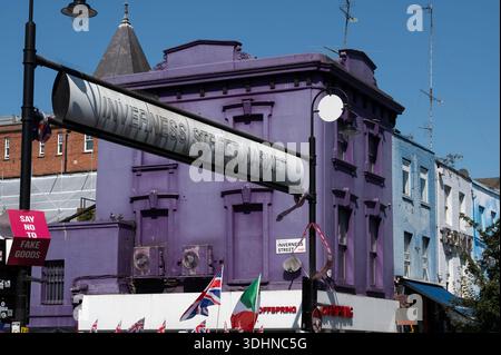 Una vivace scena serale all'Inverness Street Market di Londra presenta un grande cartello metallico sopra la testa e un edificio dipinto di viola con aria condizionata Foto Stock