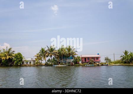 Alappuzha, India - 6 marzo 2023: Diverse case lungo il fiume con esterni colorati e palme circostanti sono visibili lungo le acque secondarie sotto un cielo limpido. Foto Stock