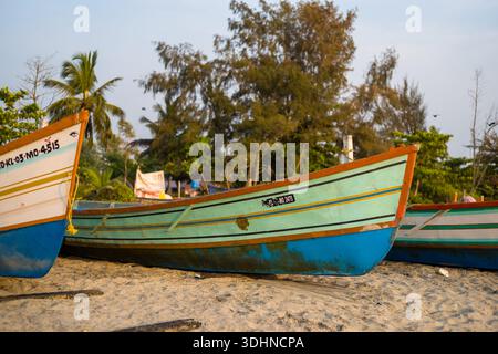 Alappuzha, India - 5 marzo 2023: Vista ravvicinata di diverse colorate barche da pesca in legno che riposano su una spiaggia sabbiosa, con alberi e vegetazione sullo sfondo sotto un cielo limpido. Foto Stock