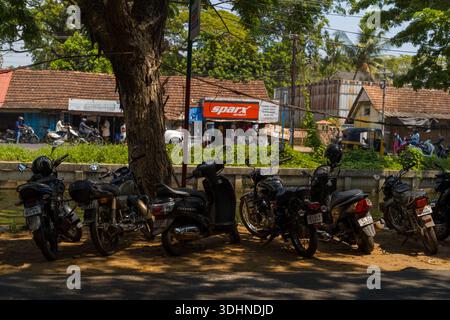 Alappuzha, India - 7 marzo 2023: Diverse motociclette e scooter sono parcheggiate sotto un grande albero accanto a un canale, con edifici tradizionali con tetto piastrellato, cartelli per negozi e persone visibili dall'altra parte dell'acqua. Foto Stock