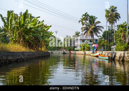 Alappuzha, India - 7 marzo 2023: Un canale delimitato da argini di pietra con piante di banane, palme, una casa residenziale e piccole barche ormeggiate lungo l'acqua, con una persona in piedi vicino alle barche sotto un cielo limpido. Foto Stock