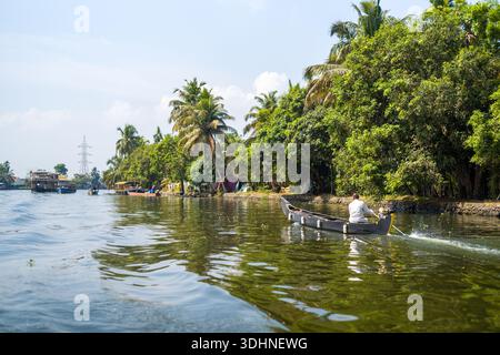 Alappuzha, India - 6 marzo 2023: Si vede un uomo guidare una piccola barca in legno motorizzata lungo un canale delimitato da fitti alberi e palme, con altre barche e case galleggianti visibili in lontananza sotto un cielo limpido. Foto Stock