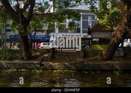 Alappuzha, India - 6 marzo 2023: I gradini conducono dal canale alla strada dove il bar del Magnet Hotel, i negozi nelle vicinanze, le motociclette parcheggiate e molte persone sono visibili sotto grandi alberi. Foto Stock