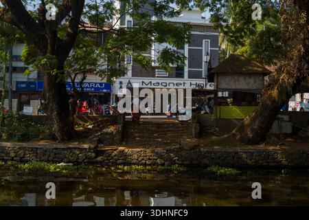Alappuzha, India - 6 marzo 2023: Si vedono persone che camminano vicino all'ingresso del bar Magnet Hotel e ai negozi adiacenti, tra cui un bancomat SBI, con motociclette parcheggiate e grandi alberi in primo piano lungo il canale. Foto Stock