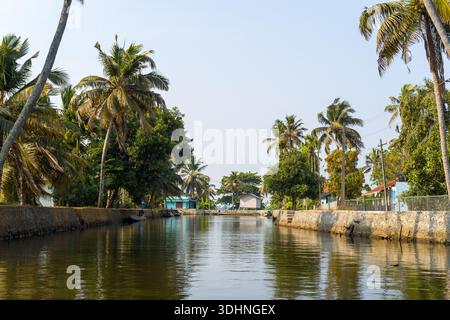 Alappuzha, India - 7 marzo 2023: Un canale delimitato da ricami in pietra è fiancheggiato da palme e case residenziali, con una piccola barca ormeggiata lungo il lato sinistro e cielo limpido sopra. Foto Stock