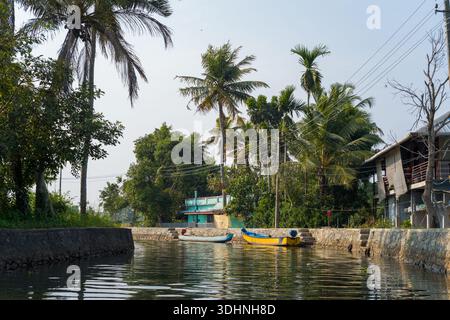 Alappuzha, India - 7 marzo 2023: Un canale delimitato da argini in pietra con palme, linee elettriche, case residenziali e due piccole barche ormeggiate lungo l'acqua sotto un cielo limpido. Foto Stock