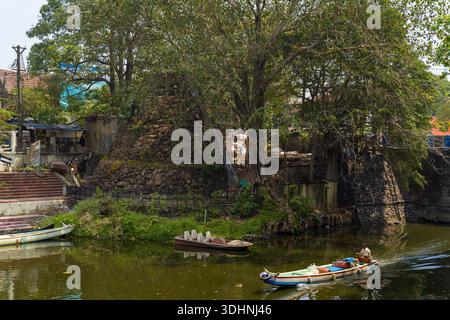 Alappuzha, India - 7 marzo 2023: Una struttura in pietra con un grande albero si erge accanto ad un canale, con un uomo che regna su una piccola barca che trasporta contenitori sull'acqua e gradini che scendono verso il canale visibili sulla sinistra. Foto Stock