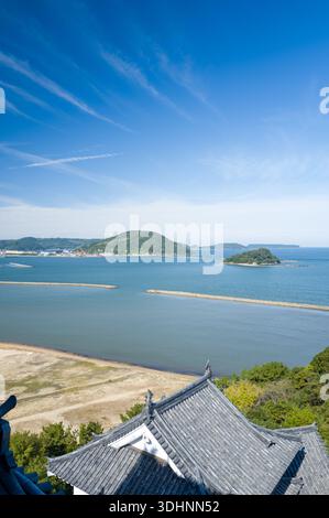 Karatsu, Giappone - 8 novembre 2024: L'immagine mostra il tradizionale tetto piastrellato del castello di Karatsu in primo piano, con una spiaggia sabbiosa, frangiflutti e diverse isole visibili nella baia sotto un cielo azzurro. Foto Stock