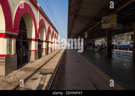 Tundla, India - 15 marzo 2024: Vista lungo una piattaforma ferroviaria presso la stazione ferroviaria di Tundla Junction, che mostra archi rossi e bianchi in stile coloniale sulla sinistra, area d'attesa coperta con segnaletica e persone sulla destra, e binari ferroviari che attraversano il centro sotto il cielo limpido. Foto Stock