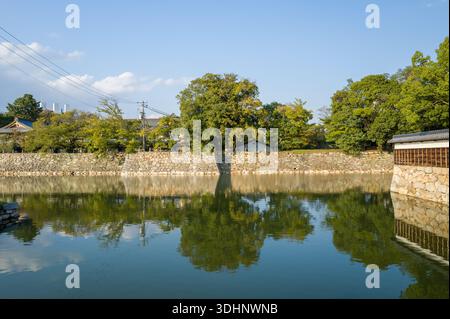 Hiroshima, Giappone - 4 novembre 2024: L'immagine mostra il muro di pietra e il fossato del castello di Hiroshima con alberi e una tradizionale struttura in legno, riflessa nell'acqua sotto un cielo azzurro. Foto Stock