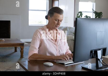 Donna anziana seduta alla scrivania di legno a casa, digitando sulla tastiera, guardando il monitor, indossando il rosa Foto Stock