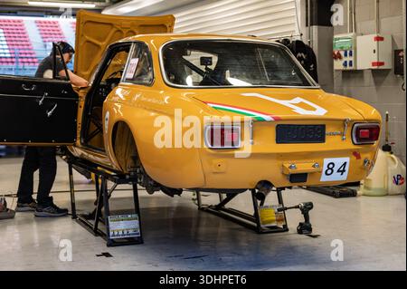 Vista posteriore di una classica Alfa Romeo 1750 GTAm 1971 arancione-giallo in un garage da corsa, rialzata su martinetti con cofano aperto, un meccanico che esegue riparazioni Foto Stock