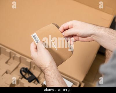Primo piano di mani maschili aprendo delicatamente una guaina di cartone marrone, eventualmente contenente manuali o accessori, parte di una nuova spedizione di acquisto online e. Foto Stock