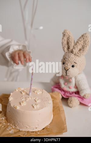 La mano di un bambino raggiunge una piccola torta di compleanno con "1 anno" scritto in glassa, accompagnato da un coniglietto a maglia in un vestito rosa, messo contro una m. Foto Stock