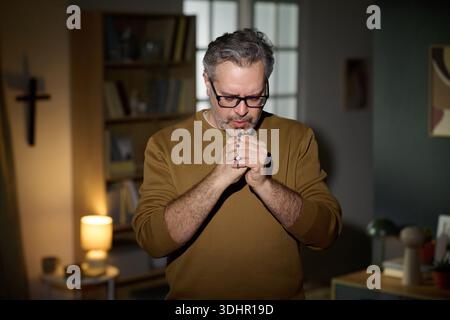Uomo di mezza età che prega in casa tenendo il Rosario con gli occhi chiusi Foto Stock