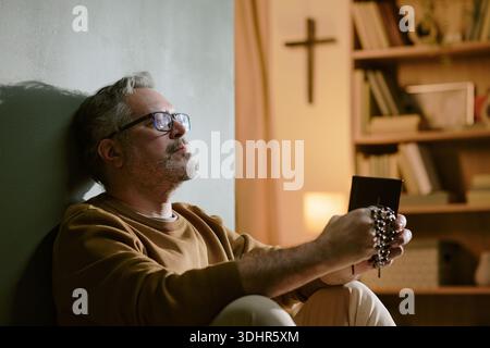 Uomo di mezza età che prega con perline di Rosario seduti al chiuso Foto Stock