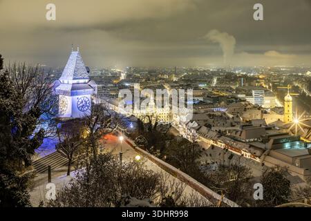 Vista della torre dell'orologio illuminata che si erge in alto nel paesaggio innevato, con le luci della città che brillano sotto a Graz, Stiria, Austria. Foto Stock