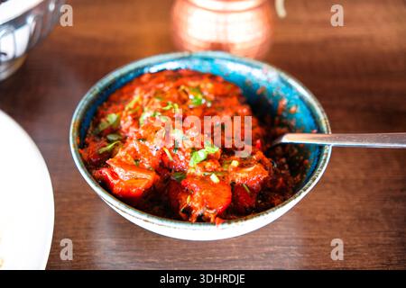 Primo piano di curry rosso indiano servito in una ciotola di metallo su un tavolo di legno, con una ricca salsa a base di pomodoro, contorno alle erbe e tavolo tradizionale Foto Stock