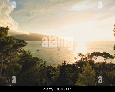 Tramonto panoramico su un mare tranquillo con barche a vela e lussureggiante costa in primo piano. Elba Italia Foto Stock
