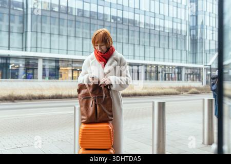 Donna di mezza età che usa lo smartphone con i bagagli in città Foto Stock