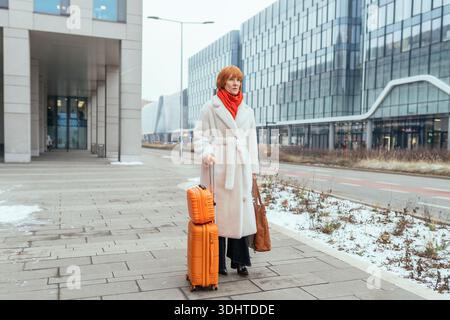 Donna matura a tutto il corpo con valigia arancione che cammina vicino a moderni edifici di uffici Foto Stock