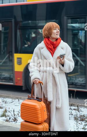 Donna caucasica premurosa con i capelli rossi che usa lo smartphone vicino all'autobus cittadino Foto Stock