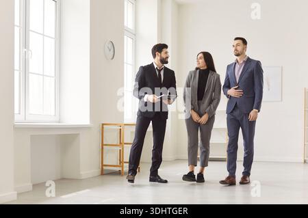 Agente immobiliare e coppia di clienti in camera vuota, uomini d'affari in piedi, guardarsi intorno Foto Stock