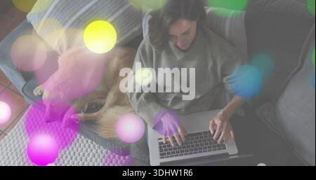Donna che scrive in maglione grigio e pantaloni scuri sul divano di casa, con laptop e cane Foto Stock