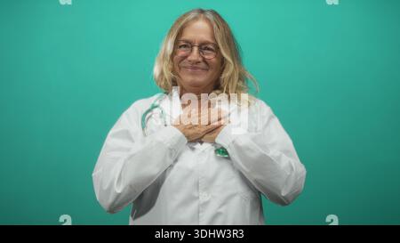Medico donna anziana che stringe le mani al petto sorridendo e guardando in alto in uno sfondo di tè dello studio; gioia gratitudine compassione. Foto Stock