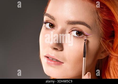 La giovane donna dai capelli arancioni applica l'eyeliner colorato, mostrando il suo fascino artistico. Foto Stock